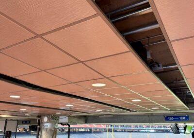 Removed ceiling panels exposing pipes and ducts in an indoor stadium concourse, with empty bleachers visible beyond the opening