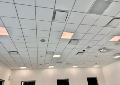 Drop-ceiling classroom with square tiles, recessed lights, vents, and a ceiling-mounted projector; white walls and three dark windows along the far wall.