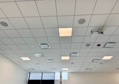 Ceiling grid with square tiles, recessed lights, vents, and a ceiling-mounted projector; windows along the far wall reveal natural light.