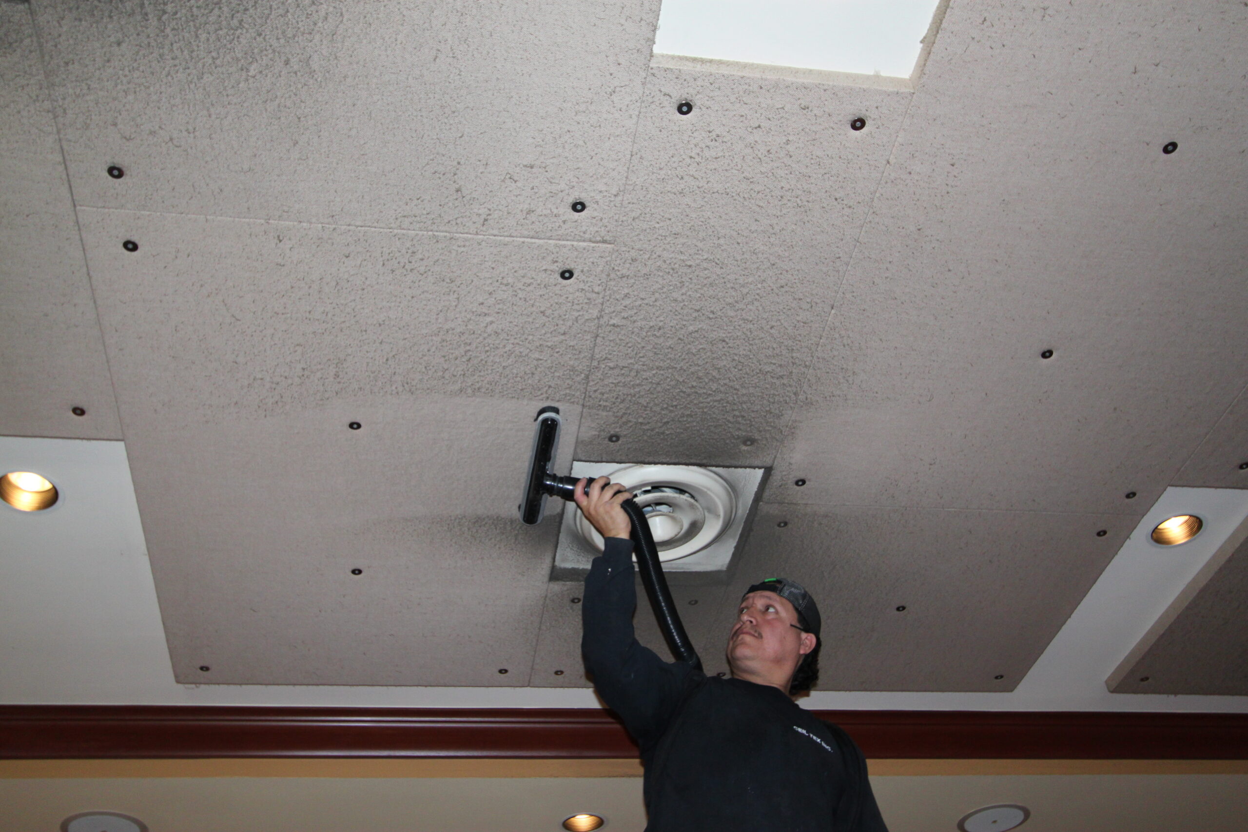 Worker vacuuming or cleaning a circular ceiling vent while installing gray drywall panels on the ceiling.